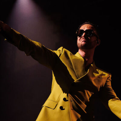 C. Tangana, El Madrileño, en el escenario, vestido con un traje amarillo brillante y gafas de sol, mientras sostiene un micrófono bajo una luz de foco dramática.