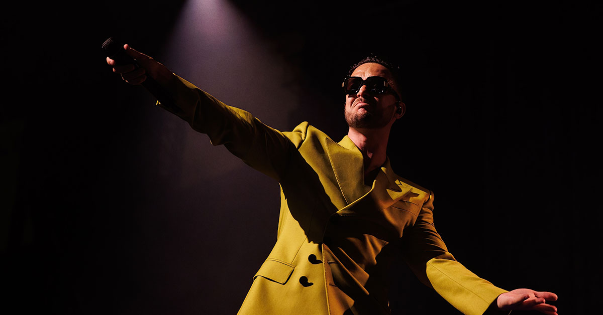 C. Tangana, El Madrileño, en el escenario, vestido con un traje amarillo brillante y gafas de sol, mientras sostiene un micrófono bajo una luz de foco dramática.